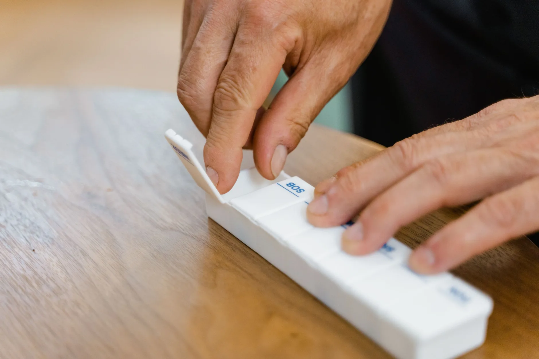 An older person&rsquo;s hands filling a weekly pill organizer on a wooden table