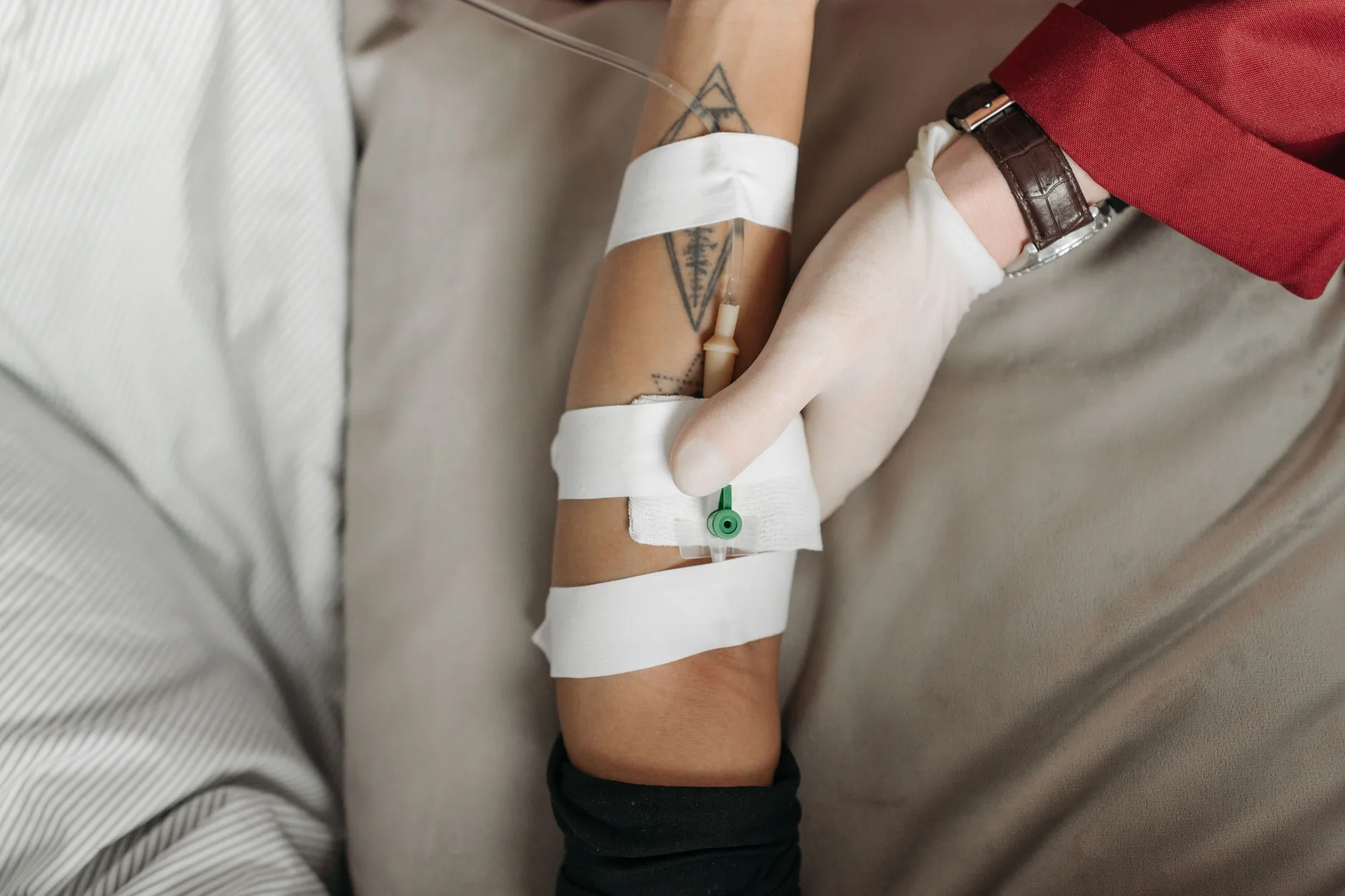 Une soignante gant&eacute;e maintenant une perfusion intraveineuse fix&eacute;e &agrave; l&rsquo;avant-bras d&rsquo;un patient